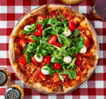 A Neapolitan-style pizza with fresh arugula, mozzarella, and cherry tomato halves, plus a glass of red wine, and shakers with red pepper flakes and Parmesan cheese on a red and white checkered tablecloth  